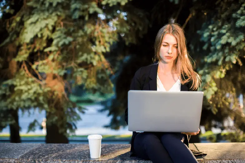 A woman sitting on a wall outdoors, working on a laptop.